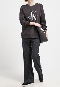 Felpa nera con grande logo bianco "CK", abbinata a pantaloni larghi a righe sottili scuri e scarpe slip-on nere, che tiene una borsa nera.