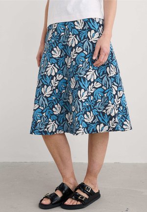 Femme portant une jupe mi-longue avec un motif de feuilles bleu et blanc, un haut blanc et des sandales noires à brides bouclées, debout sur un sol gris.