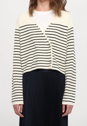 Femme portant un cardigan blanc avec des rayures horizontales noires et des boutons argentés par-dessus un top blanc et une jupe plissée noire.