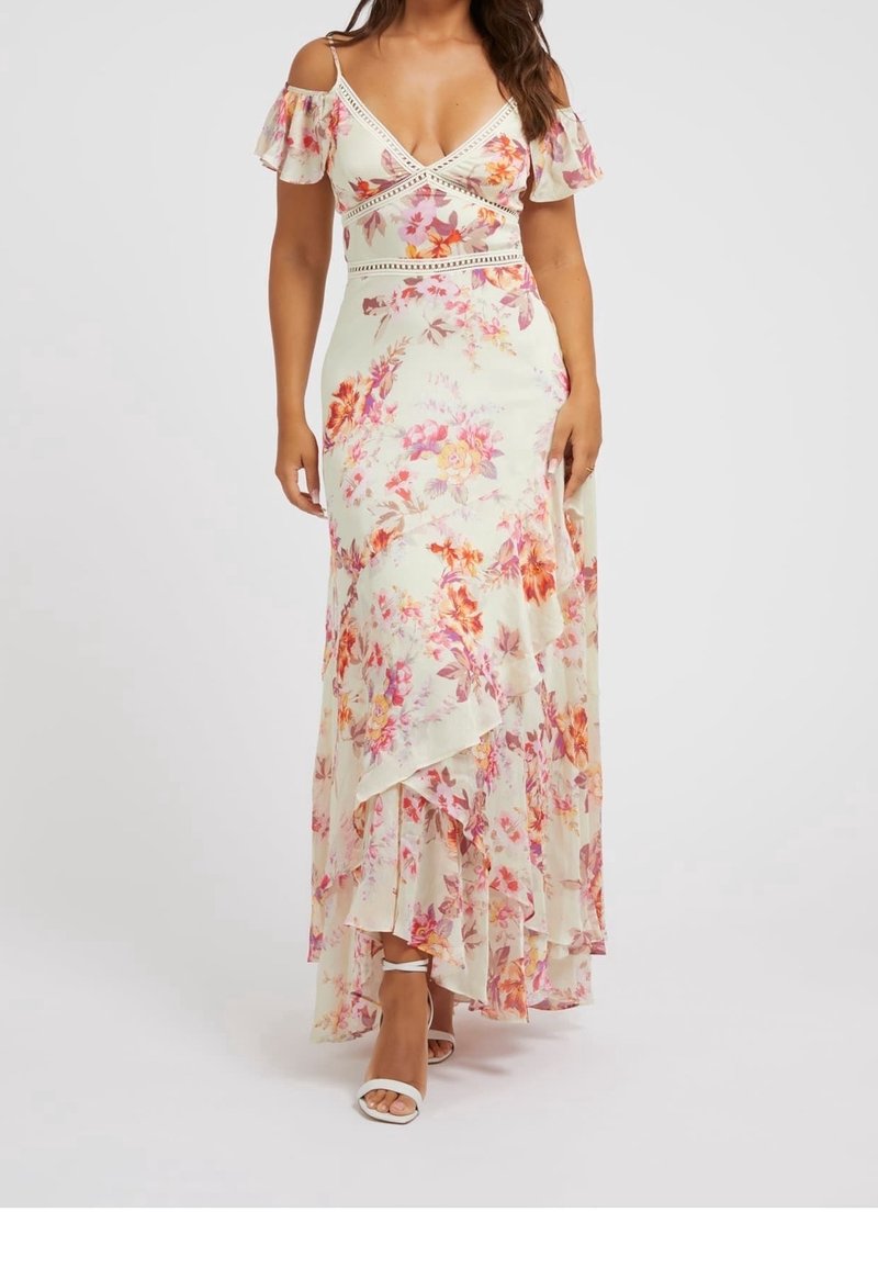Femme portant une robe maxi blanche à fleurs, épaules dénudées, avec des volants, et des talons blancs à bride cheville, se tenant devant un fond uni.