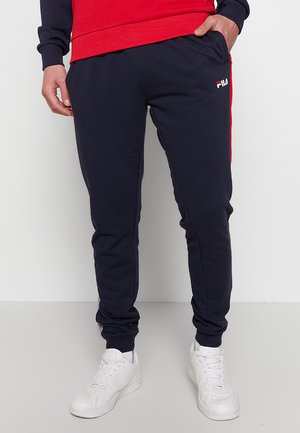 Persona che indossa pantaloni della tuta FILA blu navy con strisce laterali rosse, scarpe da ginnastica bianche e una felpa rossa e blu navy, in piedi davanti a uno sfondo semplice.