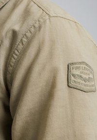 Beige bomulds skjorte med struktureret stof, synlige sømme og et patch med "PINE LEGEND AVIATOR" samt vingedesign på skulderen.