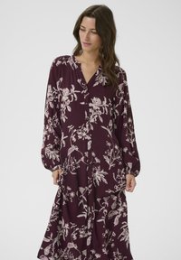 Modell in einem langen, burgunderfarbenen Blumenkleid mit Knopfdetails, der ein sanftes Lächeln zeigt und in einer entspannten Pose vor einem neutralen Hintergrund steht.