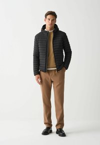 Chaqueta acolchada negra con capucha, que cuenta con cierre de cremallera y diseño acolchado, llevada sobre un suéter beige y combinada con pantalones marrones.