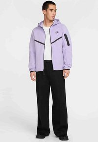 Giacca viola con cappuccio e dettagli neri, dotata di tasca con zip, abbinata a pantaloni neri morbidi. Texture liscia, design casual, stile sportivo.
