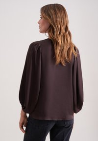 Blusa marrone a maniche lunghe con una texture liscia, scollatura rotonda e dettagli arricciati sulle spalle. Indossata con pantaloni in denim scuro.