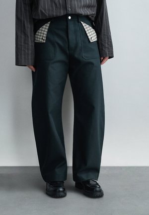 Osoba nosiaca čierne široké nohavice s viditeľnou šedou károvanou výstelkou vrecka, čierne masívne topánky a tmavú pruhovanú košeľu s dlhými rukávmi.