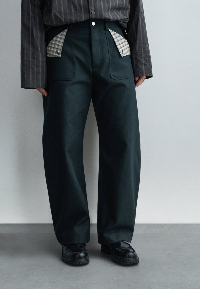 Άτομο που φοράει μαύρο παντελόνι με πλατιά μπατζάκια και ορατές γκρι καρό επενδύσεις στις τσέπες, μαύρα chunky παπούτσια και σκούρο ριγέ πουκάμισο με μακριά μανίκια.