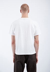 T-shirt blanc à manches courtes en coton, avec un col rond et une coupe droite, porté avec un jean marron foncé. Détails de couture visibles.