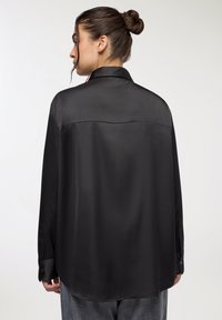 Camicia nera in satin con una consistenza liscia, maniche lunghe e un colletto classico. Presenta un dettaglio a yoke sulla schiena e polsini abbottonati.