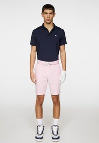 Laivastonsininen polo-paita, vaaleanpunaiset shortsit, valkoiset käsineet ja siniset korkeavartiset tennarit valkoisilla yksityiskohdilla. Polo-paidassa on logo ja metallinen vyönsolki.