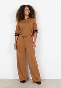 Mujer con cabello rizado usando un conjunto marrón de dos piezas con ribete negro, de pie y sonriendo frente a una pared blanca lisa.