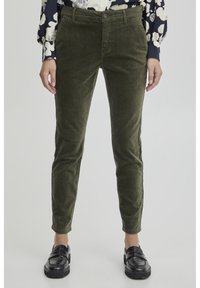 Pantaloni in corduroy verde oliva con vestibilità slim, dotati di chiusura a bottone, tasche frontali e tessuto testurizzato. Indossati con una blusa a fantasia e mocassini neri.
