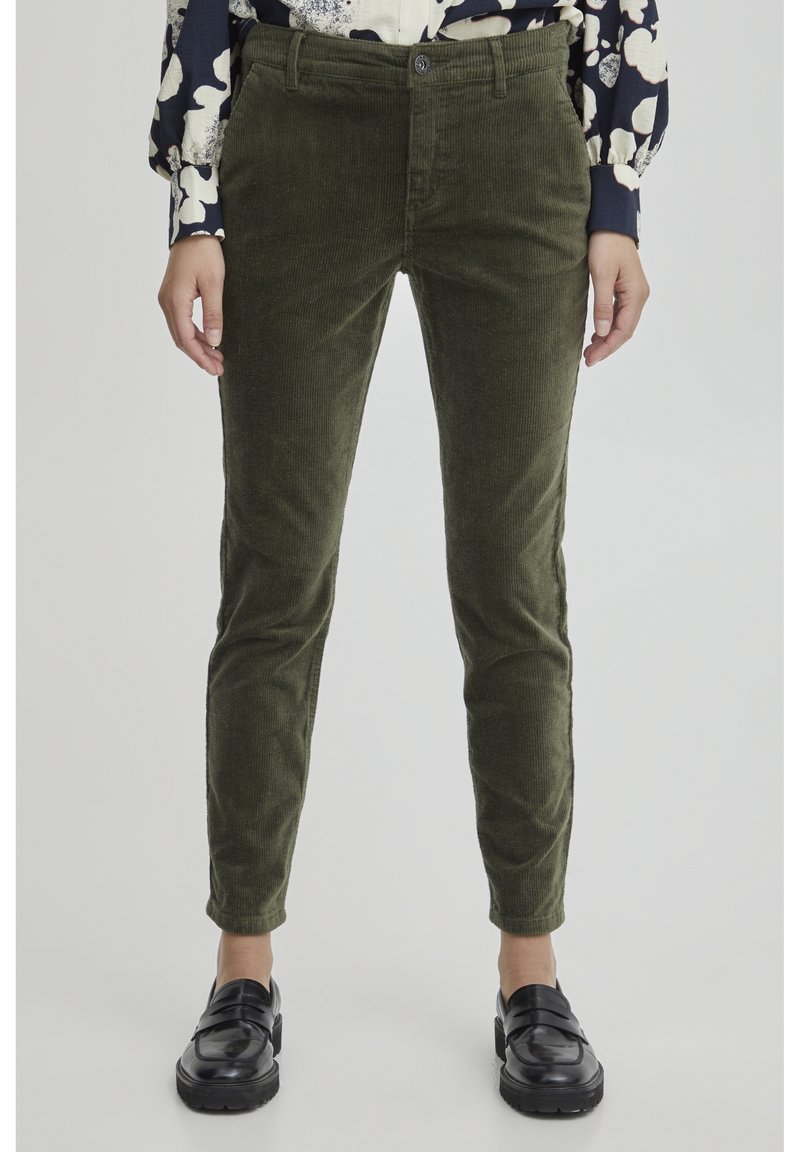 Pantaloni in corduroy verde oliva con vestibilità slim, dotati di chiusura a bottone, tasche frontali e tessuto testurizzato. Indossati con una blusa a fantasia e mocassini neri.