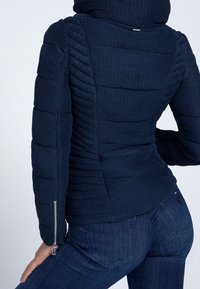 Tmavě modrá péřová bunda s texturovanou látkou, přiléhavým střihem, šikmými švy a zipy na manžetách.