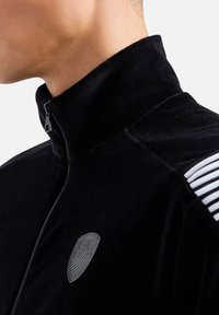 Giacca in velluto nero con zip, caratterizzata da un alto colletto, un logo tono su tono sul petto e accenti a strisce bianche sulle spalle.