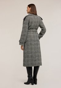 Cappotto lungo a quadri in tessuto bianco e nero, con vita tuttavia, spalle strutturate e bottoni oversized. Indossato con stivali neri al ginocchio.