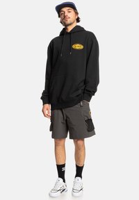 Zwarte hoodie met een geel logo, gecombineerd met grijze cargo shorts. Witte sneakers met zwarte accenten en crew sokken.