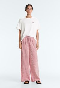 Femme debout portant un t-shirt blanc ample avec un petit motif de rose et un pantalon large à rayures roses avec des sandales noires.