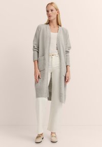 Helle graue, lange Strickjacke aus strukturiertem Maschenmaterial, mit gerippten Bündchen, zwei vorderen Taschen und offenem Design. Kombiniert über einem weißen Oberteil und Hose.