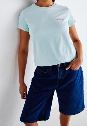 Personne portant un t-shirt Levi's bleu clair et un short en denim bleu foncé jusqu'aux genoux, une main dans la poche, debout contre un mur blanc.