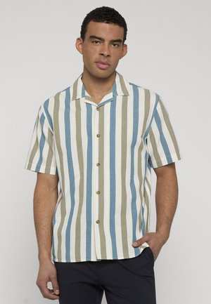 Mand iført en kortærmet skjorte med knapper og lodrette blå, beige og hvide striber, stående med den ene hånd i lommen.