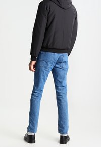 Homme portant un jean bleu, une veste noire, des chaussettes blanches et des chaussures noires, debout de dos sur un fond blanc.