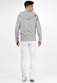 Sweatshirt à capuche gris en tissu doux, associé à un jean blanc slim-fit. Le jean présente des coutures jaunes sur les poches arrière.