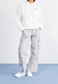 Sudadera blanca con bolsillo frontal y logo negro, pantalones cargo grises con múltiples bolsillos y zapatillas blancas con un diseño rosa.