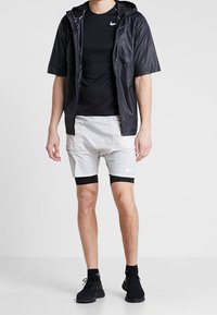 Černá krátká bunda s krátkým rukávem přes černé tričko, doplněná světle šedými šortkami a černými kompresními legínami. Celkový vzhled doplňují černé sportovní boty.