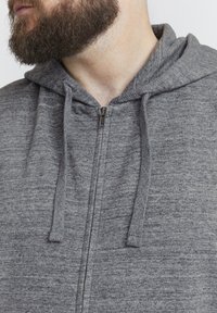 Sudadera con cremallera en color gris, hecha de un tejido texturizado, con capucha, que presenta cordones ajustables y una cremallera metálica. Sin patrones ni logotipos visibles.