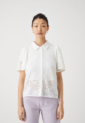 Hvit bluse i blonder med spiss krage og korte pufferm, med knapper foran og blomsterdesign, brukt over lys lavendelbukser.