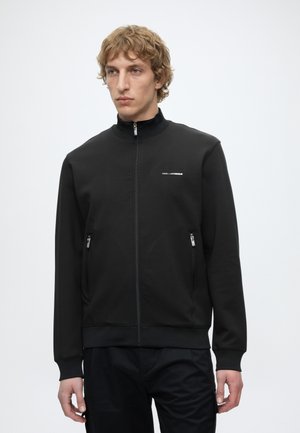 Giovane uomo con capelli biondi ricci che indossa una giacca nera con zip, tasche frontali e un logo bianco discreto sul petto.