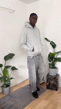 Grauer Kapuzenpullover mit Kängurutasche, hellgraue Jeans, schwarze Schuhe, stehend auf einem grauen Teppich mit grünen Pflanzen in grauen und schwarzen Töpfen.