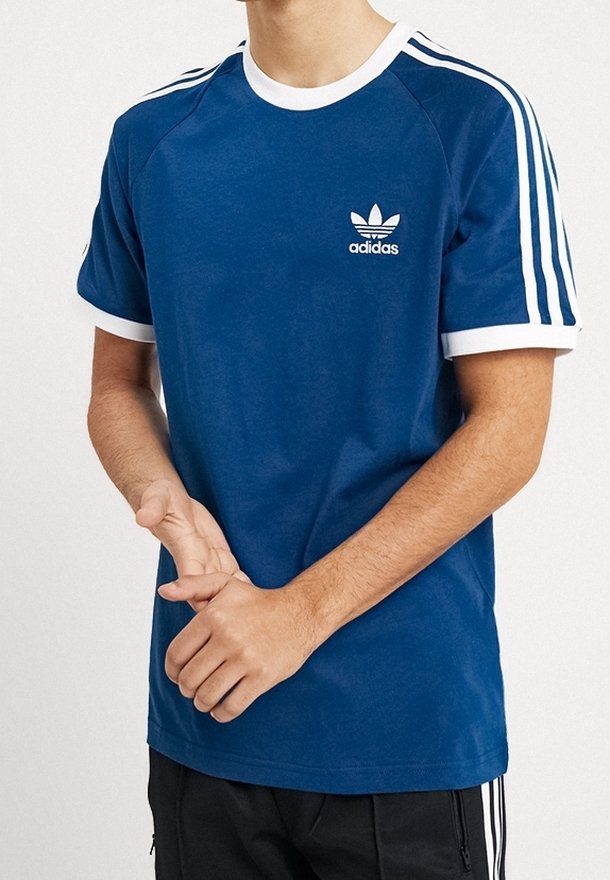 Sininen puuvillapaita, jossa on valkoinen kaulus ja hihansuut. Paitassa on valkoinen kolmiraita-kuviointi hartioilla ja pieni Adidas-logo rinnassa.