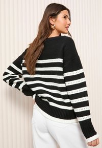 Maglione a righe bianche e nere con vestibilità ampia, polsini e orlo a coste, e dettagli ad occhiello. La vista posteriore mostra la spalla e la texture.
