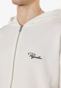 White zip-up hoodie made of ribbed fabric. Features a black embroidered "Pegadon" logo on the chest. Soft texture with a hood.