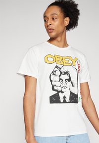 Λευκό βαμβακερό μπλουζάκι με έντονη γραφική εκτύπωση ενός γροθιά και μιας φιγούρας σε κοστούμι. Το κείμενο αναγράφει "ΥΠΑΚΟΥ" σε κίτρινο χρώμα, με κόκκινες και πράσινες λεπτομέρειες.