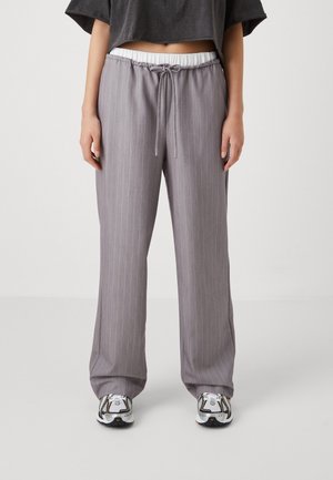 Pantalons gris à rayures verticales avec une taille élastique et un cordon de serrage. Coupe ample, se resserrant vers le bas. Portés avec des baskets blanches et noires.