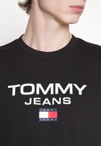 Camiseta de algodón negra con el texto en blanco "TOMMY" y "JEANS" en la parte frontal, además de un pequeño rectángulo de logo en rojo, blanco y azul.