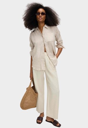 Kvinde med stort krøllet hår iført stribet beige skjorte, løse cremefarvede bukser, mørke solbriller, brune slip-on sandaler og holder en vævet taske.