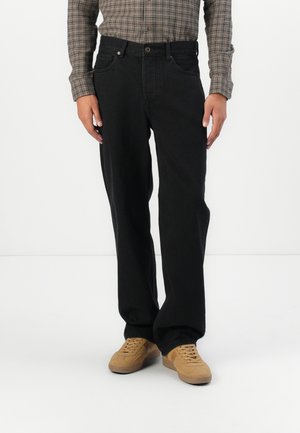 Person iført løstsiddende sorte jeans, brunternet skjorte og beige sneakers stående mod en hvid baggrund.