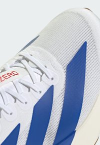 White athletic shoe with blue diagonal stripes, textured fabric upper, white laces, and a red logo on the tongue. Smooth rubber sole.