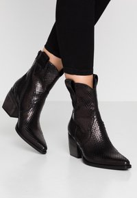 Bottines noires en simili cuir serpent, à bouts pointus, avec un talon empilé et des panneaux élastiques sur les côtés, présentant une surface texturée et des détails western.