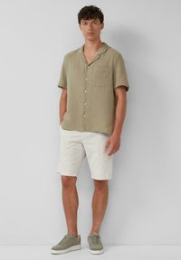 Kurzarmliges, olivgrünes Hemd mit knöpfbarem Kragen und strukturierter Stoffoberfläche, einer Brusttasche, kombiniert mit weißen Shorts und grauen Sneakers.