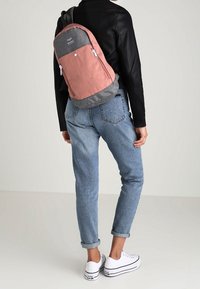 Zaino con parte anteriore in tessuto rosa e fondo grigio, dotato di due tasche con zip. Indossato da una persona con jeans arrotolati e sneakers bianche.