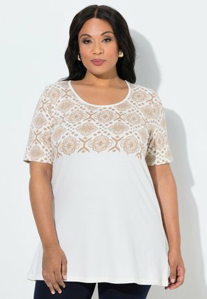 Frau trägt eine weiße Tunika mit beigen, geometrischen Mustern auf Schultern und Ärmeln, steht vor einem schlichten weißen Hintergrund.