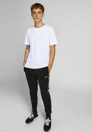 Hvid bomulds t-shirt parret med sorte joggere med grønne grafiske detaljer og sidestriber, suppleret af sorte sneakers.