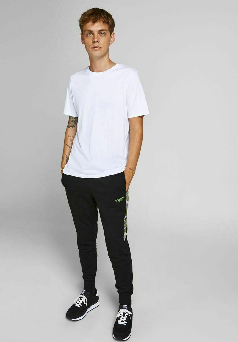 Hvid bomulds t-shirt parret med sorte joggere med grønne grafiske detaljer og sidestriber, suppleret af sorte sneakers.