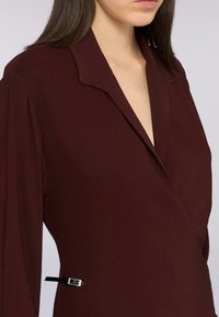 Bordeaux wikkeljurk met een inspringende kraag, lange mouwen en een zijgesp mock-up. Gemaakt van een gladde stof met een subtiele glans.